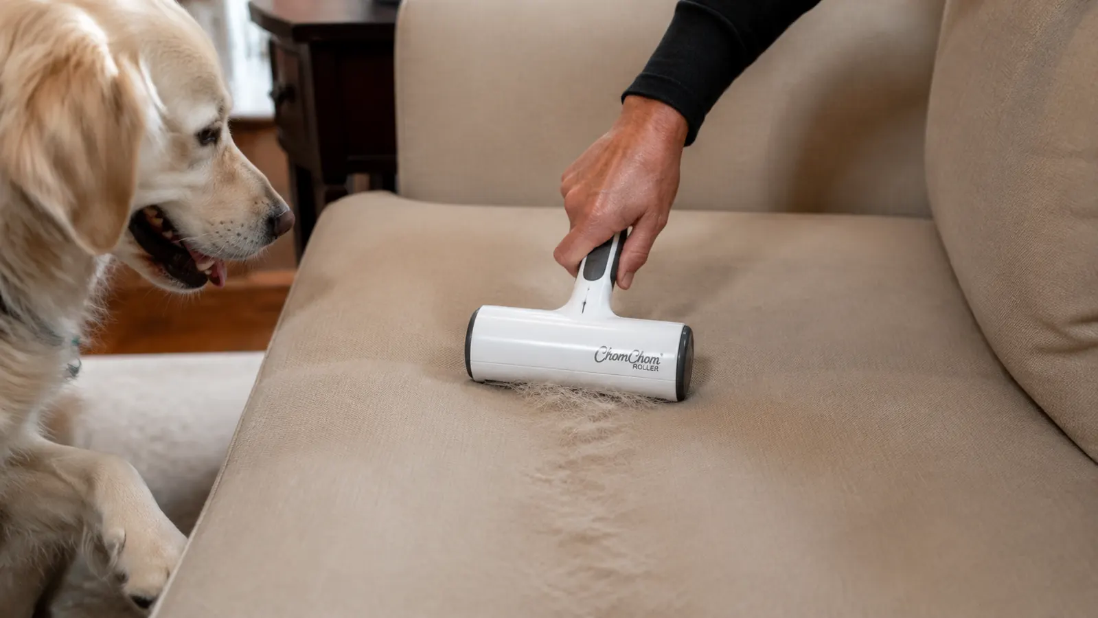 A hand pushing a handheld rolling fabric cleaner in a long straight line down the center of a beige couch cushion, leaving a clean track through the fabric